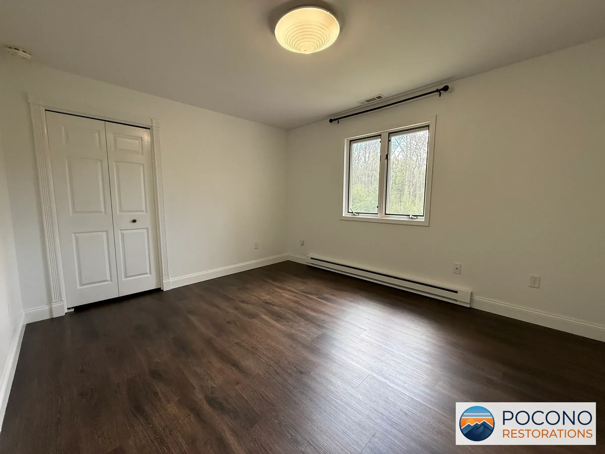 Empty bedroom with a white bi-fold closet door, dark wood-look flooring, a round ceiling light fixture, a window, and a baseboard heater.