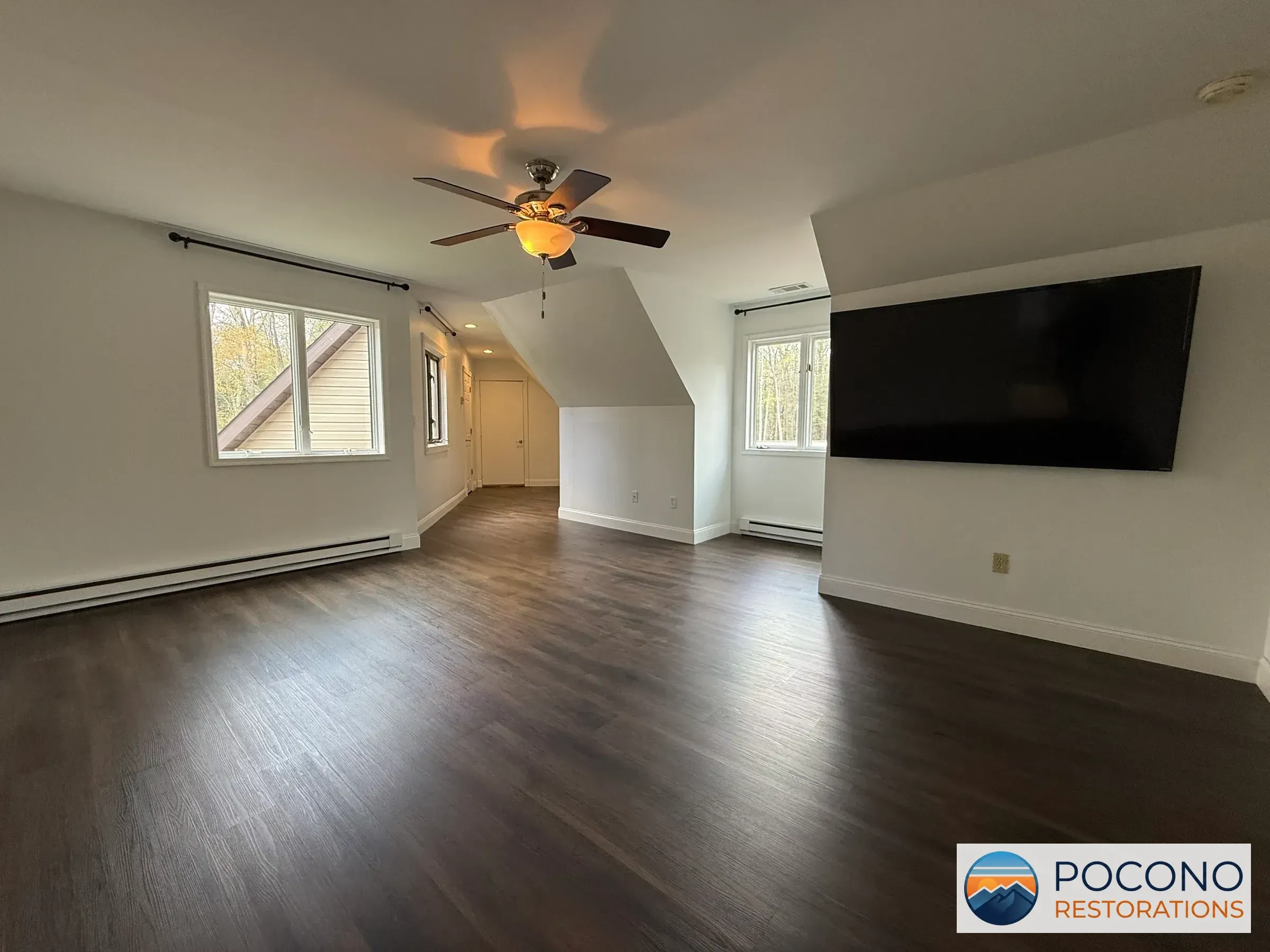 Empty upstairs room with dark wood-look plank flooring, angled ceiling, ceiling fan, wall-mounted television, and baseboard heaters below two windows.