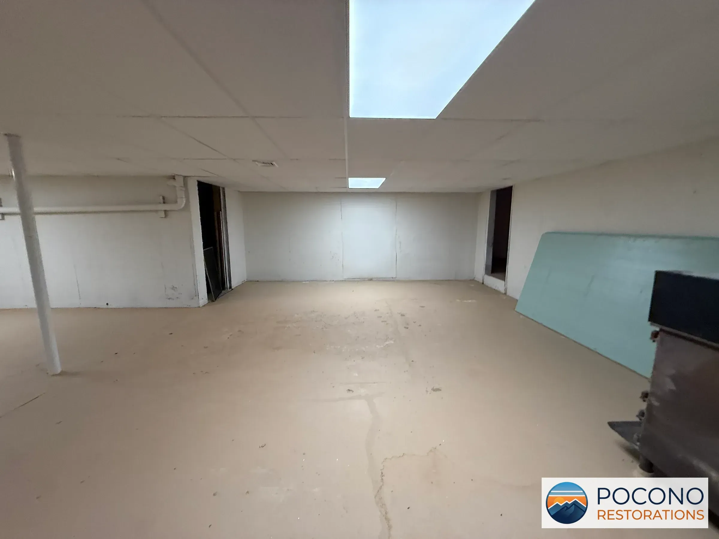 Basement corner with an illuminated ceiling panel, two door openings, a teal panel leaning against the wall, a metal equipment unit, and a stained concrete floor.