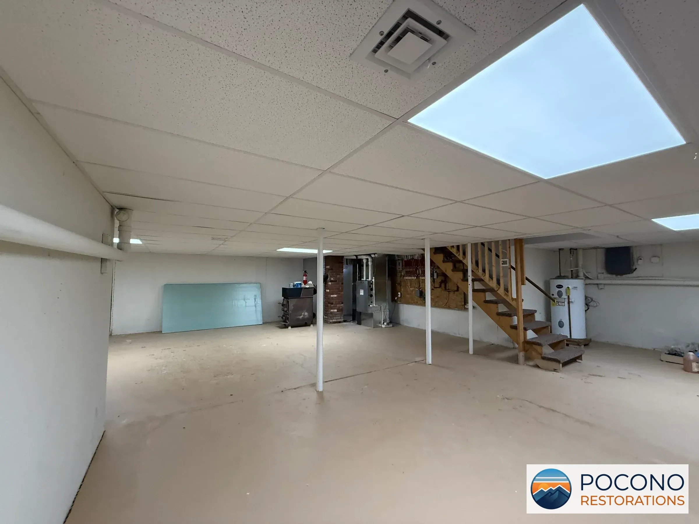 Basement space with a wooden staircase, a white water heater tank, an electrical panel, a plumbing manifold on plywood, a brick wall section, an illuminated ceiling panel, and a bare concrete floor.
