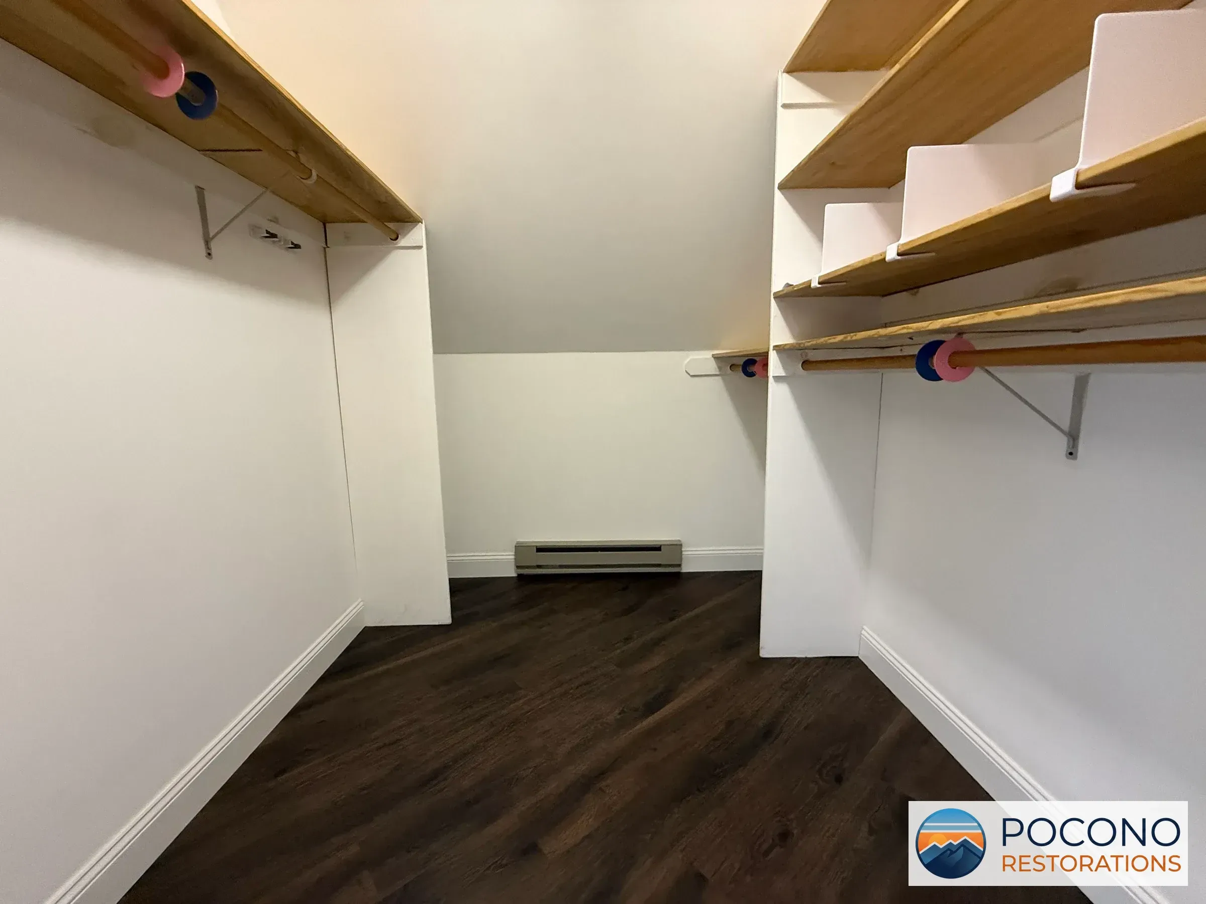 Empty walk-in closet with wood shelving and closet rods on each side, sloped ceiling, baseboard heater, and dark wood-look plank flooring.