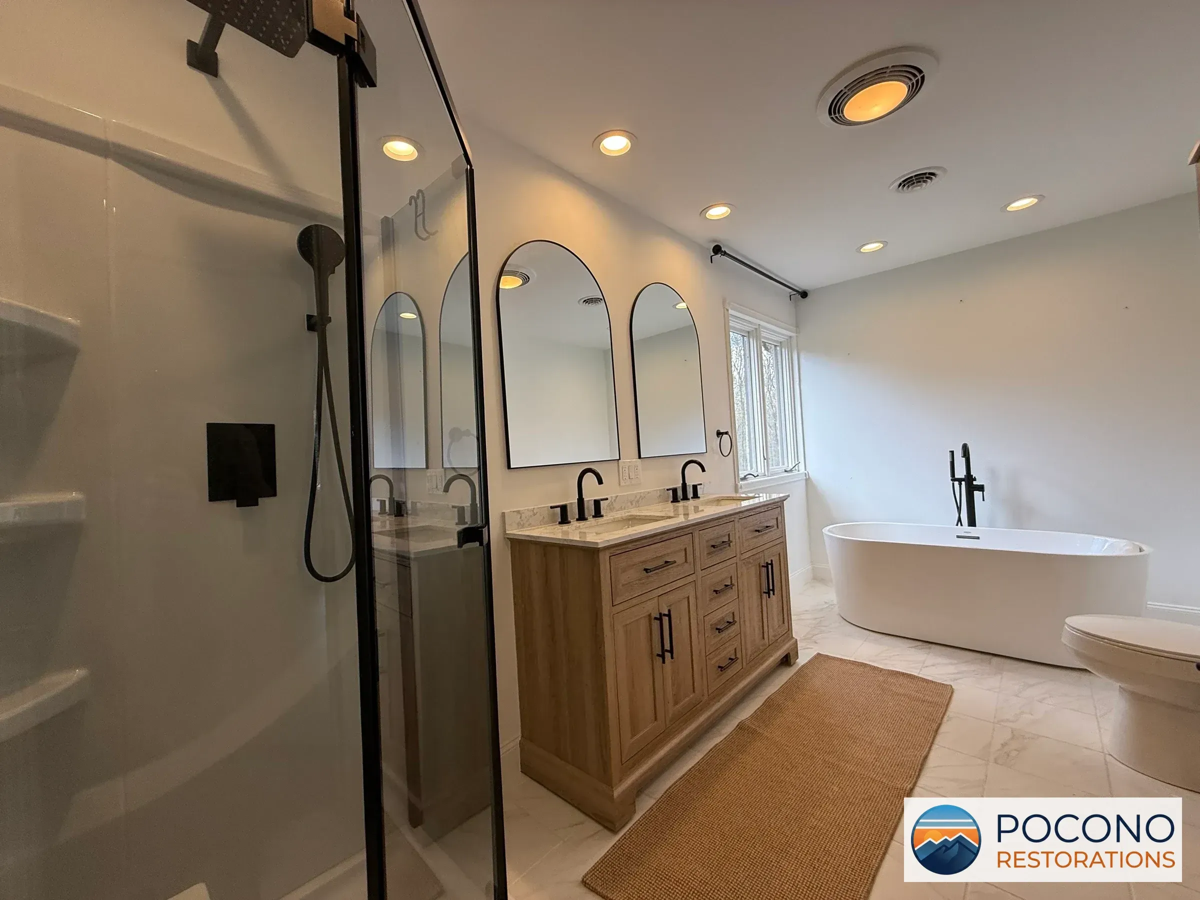 Bathroom view with glass shower enclosure, arched mirrors, wood-tone double vanity, freestanding tub, toilet, and light veined tile flooring.