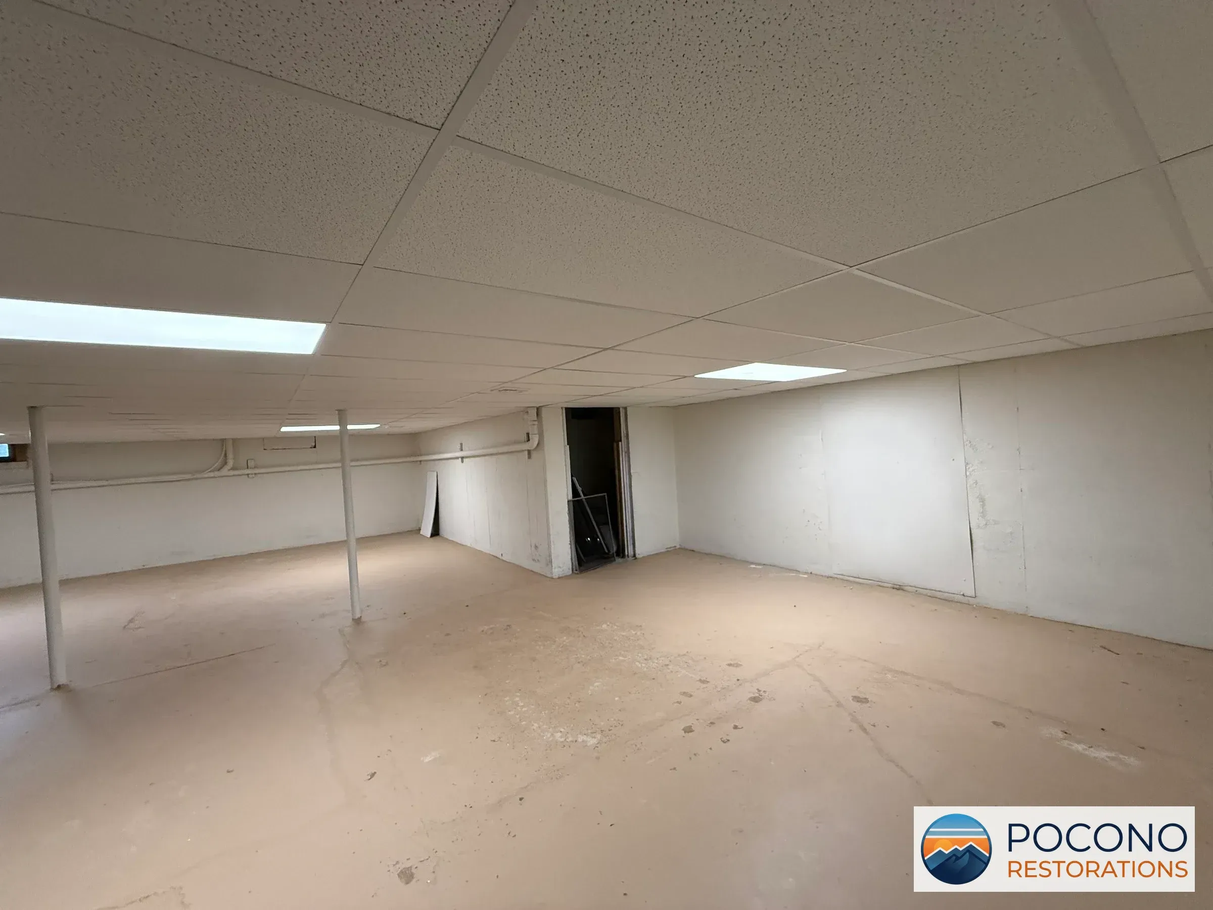 Basement area with drop ceiling tiles, one illuminated panel, a white support pole, pipes along the ceiling, a small door opening, and staining visible on the walls and floor.