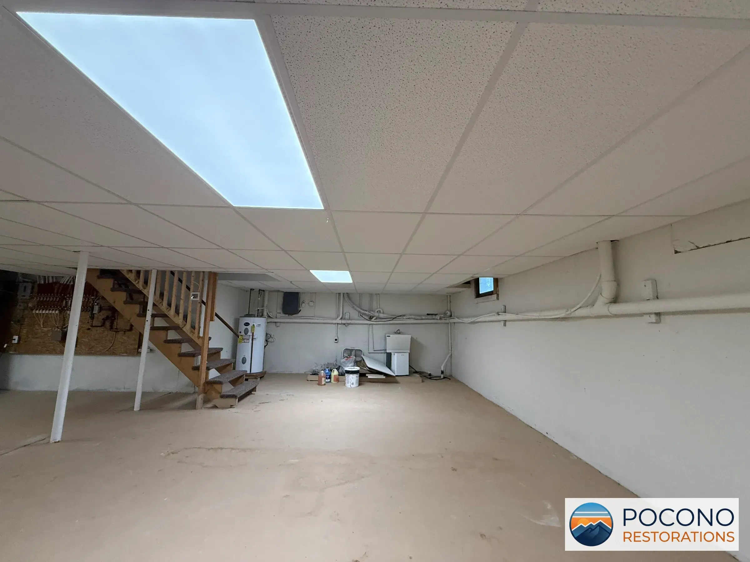 Basement with a drop ceiling, two translucent light panels, a wooden staircase, a water heater, exposed plumbing fittings, and pipes along the walls above a concrete floor.