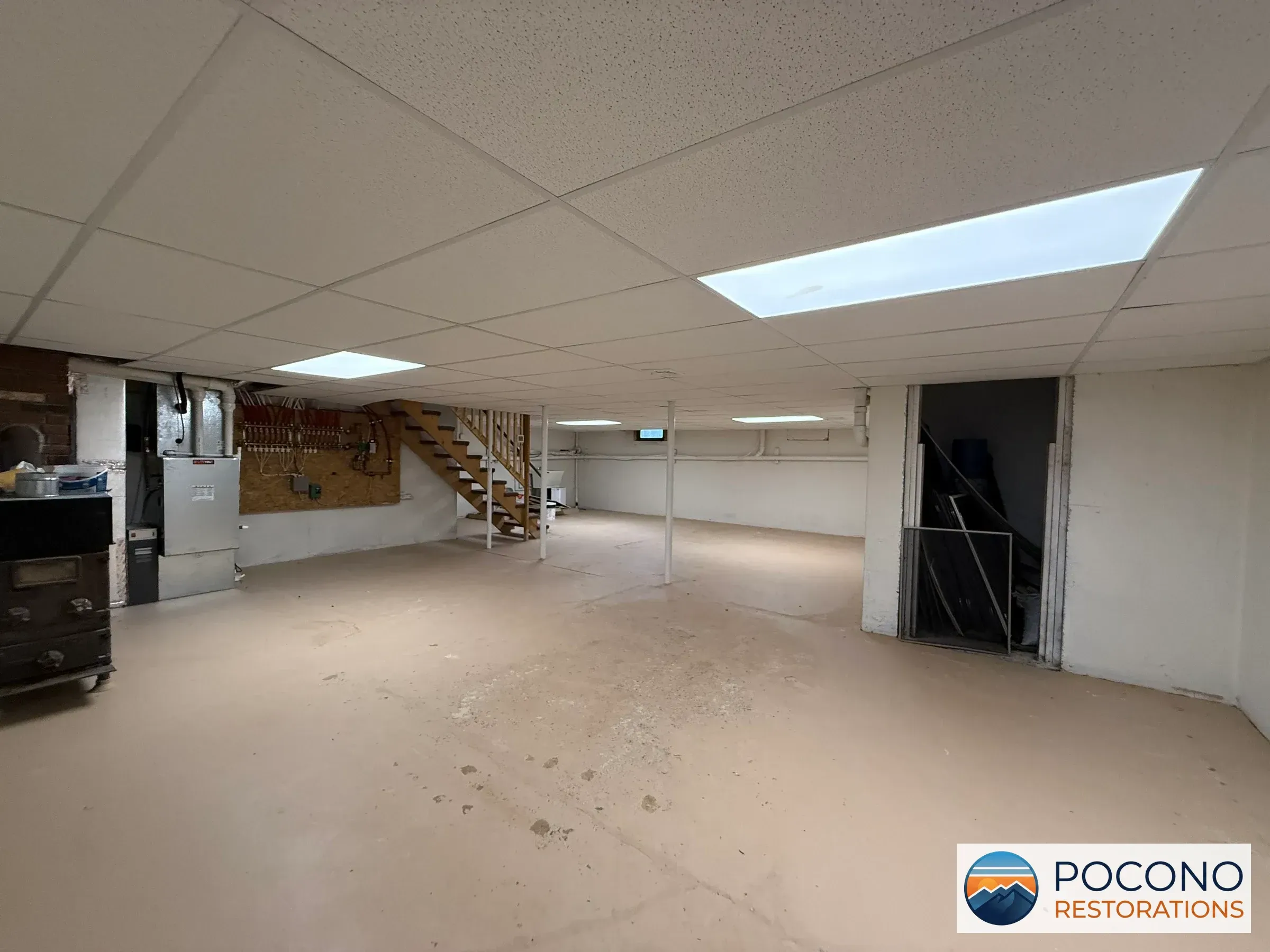 Basement with drop ceiling tiles, two illuminated ceiling panels, a wooden staircase, a support post, an electrical panel, a plumbing manifold on plywood, and a bare concrete floor; a closet doorway is visible on the right.