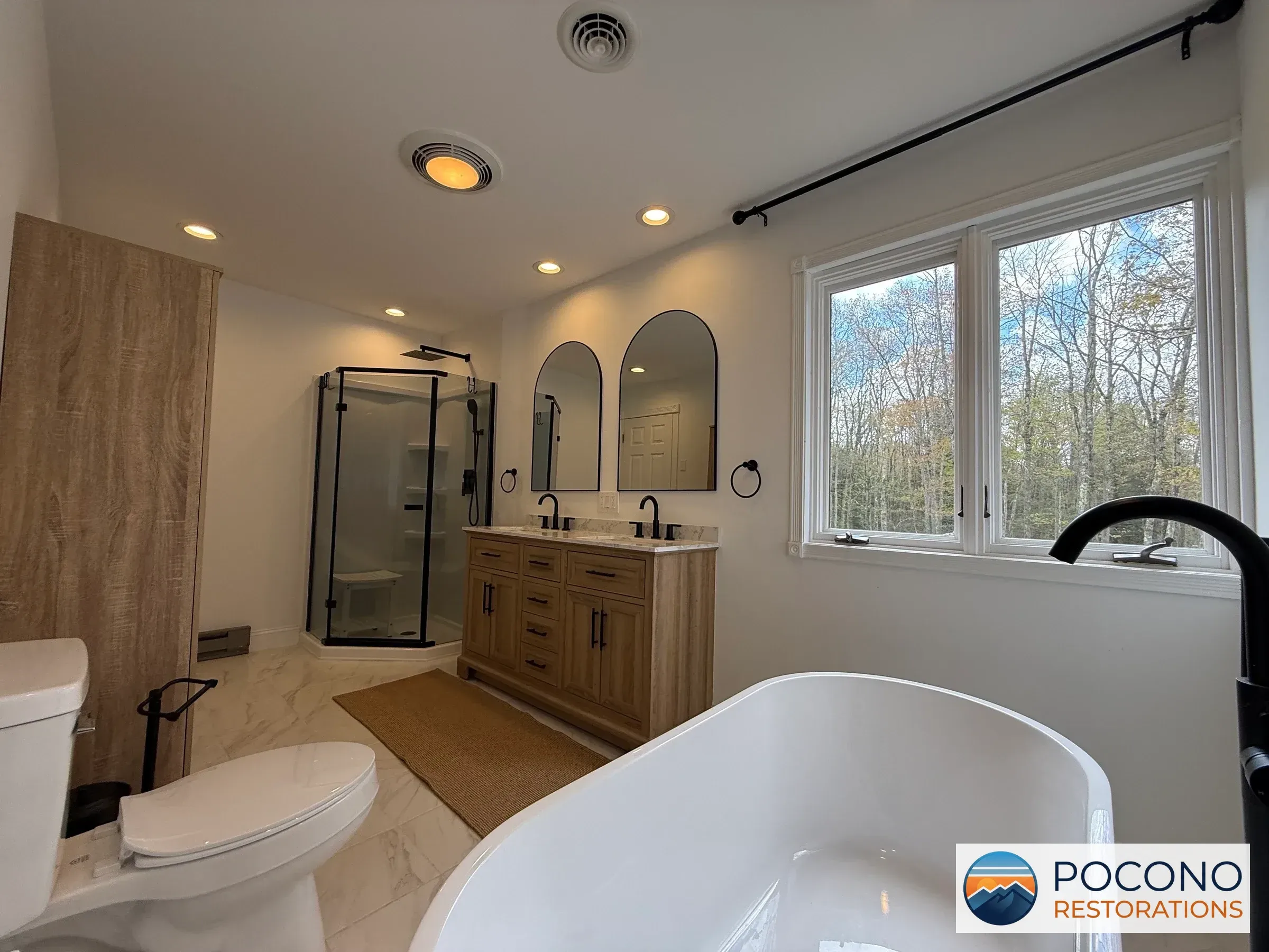 Bathroom with double wood-tone vanity, arched mirrors, corner glass shower, freestanding oval tub, toilet, and light veined tile flooring; window overlooks bare trees.
