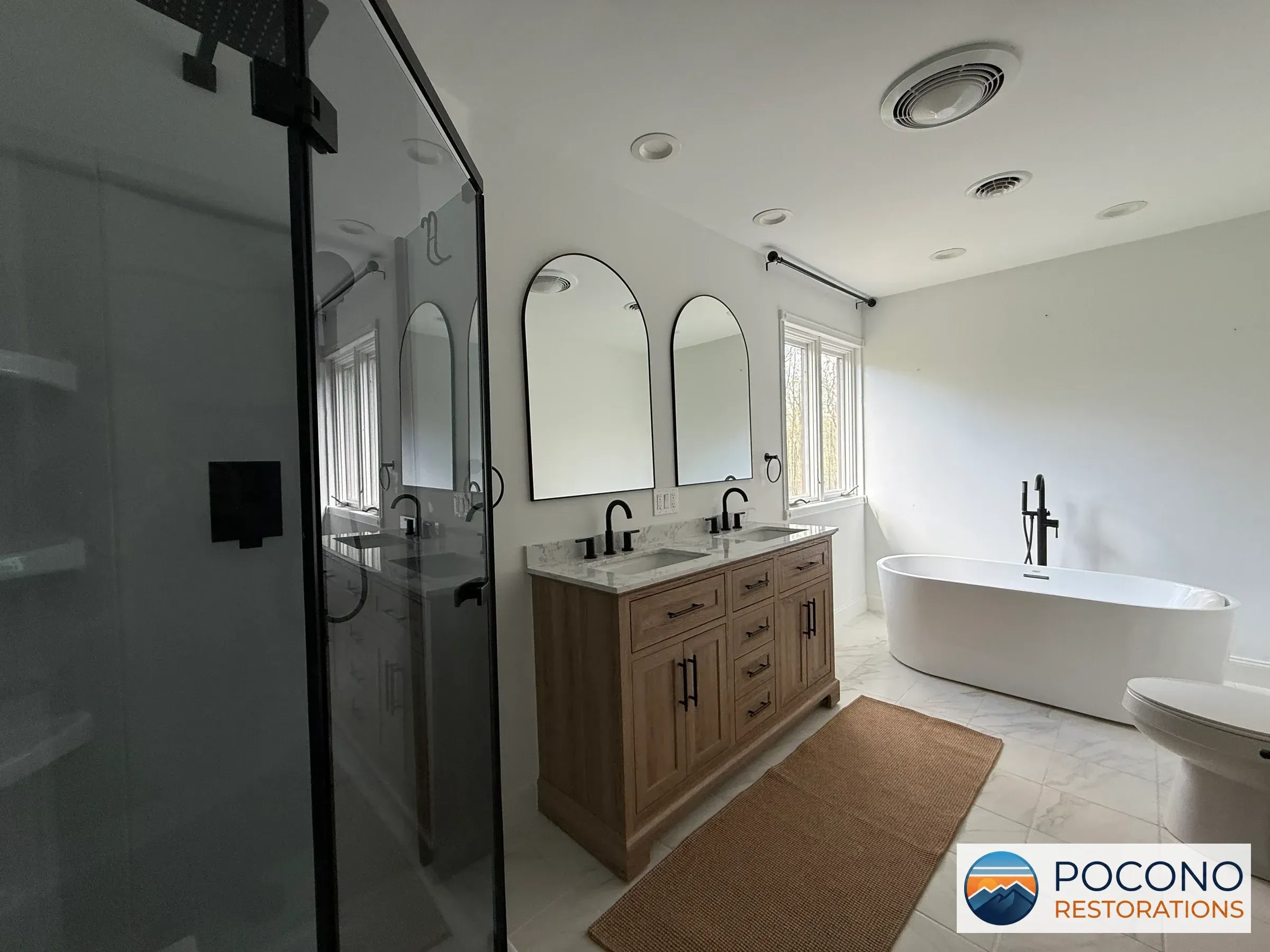 Bathroom with black-framed glass shower, wood double vanity with arched mirrors, marble-look countertop, freestanding oval tub, and tiled floor.