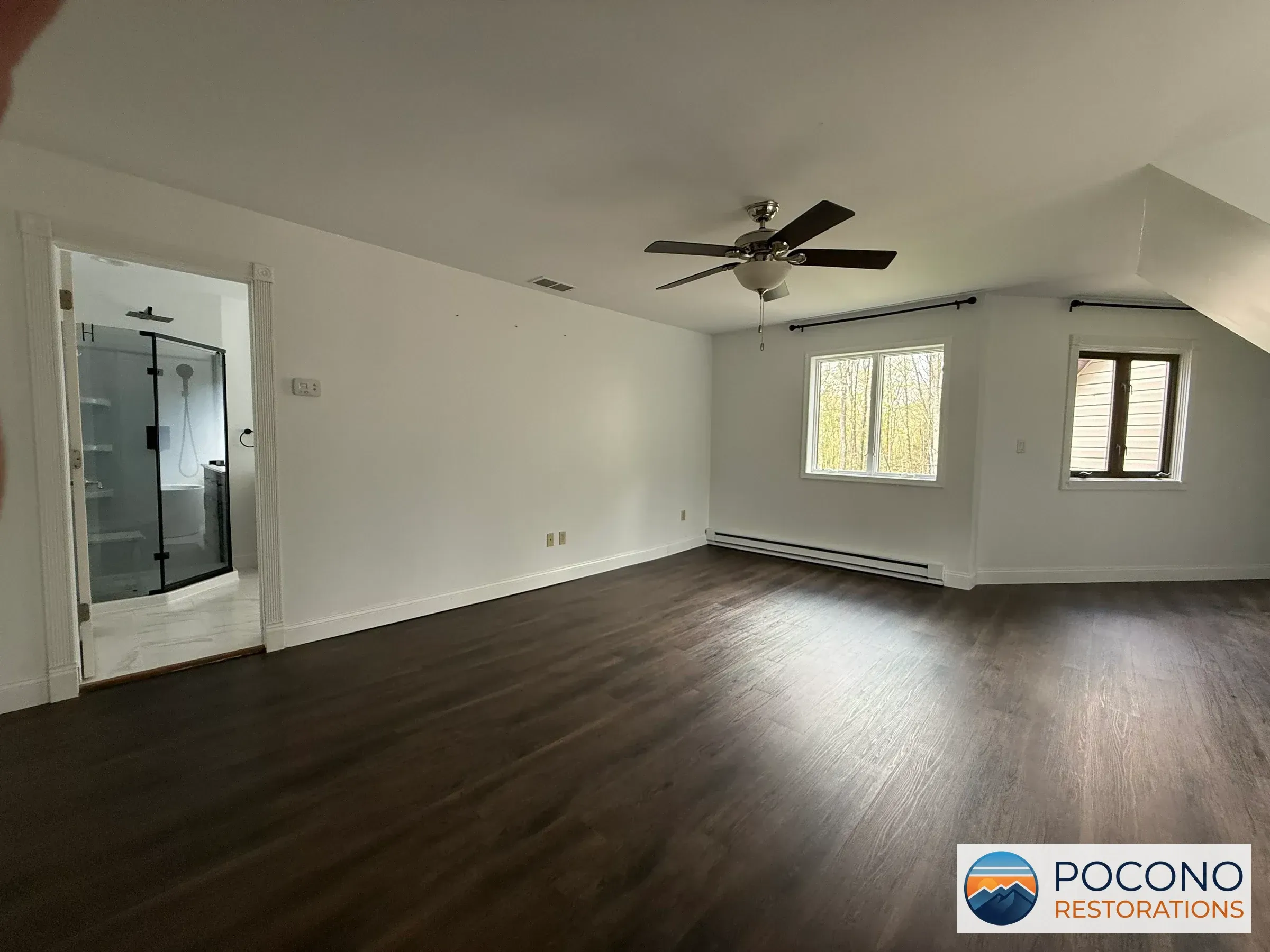 Empty upstairs bedroom with dark wood-look flooring, vaulted ceiling with a ceiling fan, two windows, and an open doorway to an attached bathroom with a glass shower.