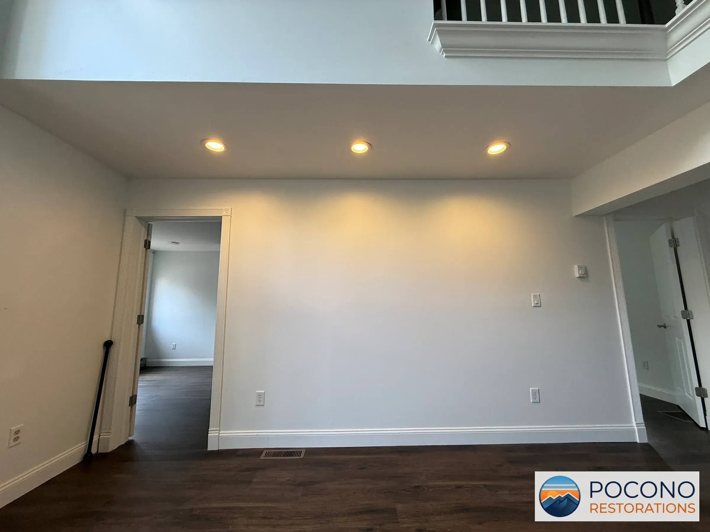 Wall corner with three recessed ceiling lights, adjoining open doorways, dark wood flooring, and a loft railing above.