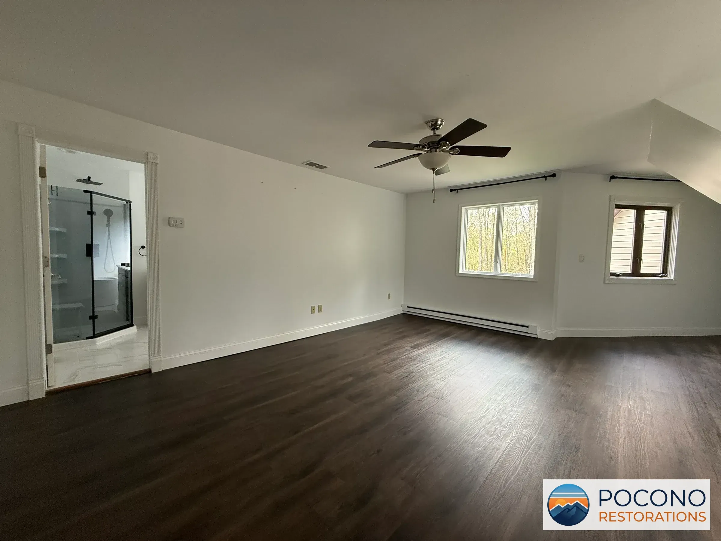 Empty upstairs room with dark wood-look flooring, ceiling fan, and an open doorway revealing a tiled bathroom with a glass shower enclosure.