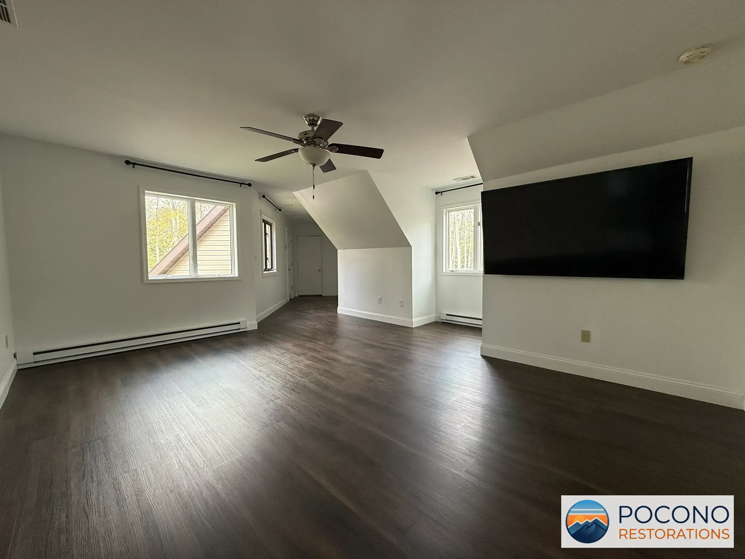 Empty upstairs room with dark wood-look flooring, ceiling fan, and a large wall-mounted TV opposite windows under a sloped ceiling.
