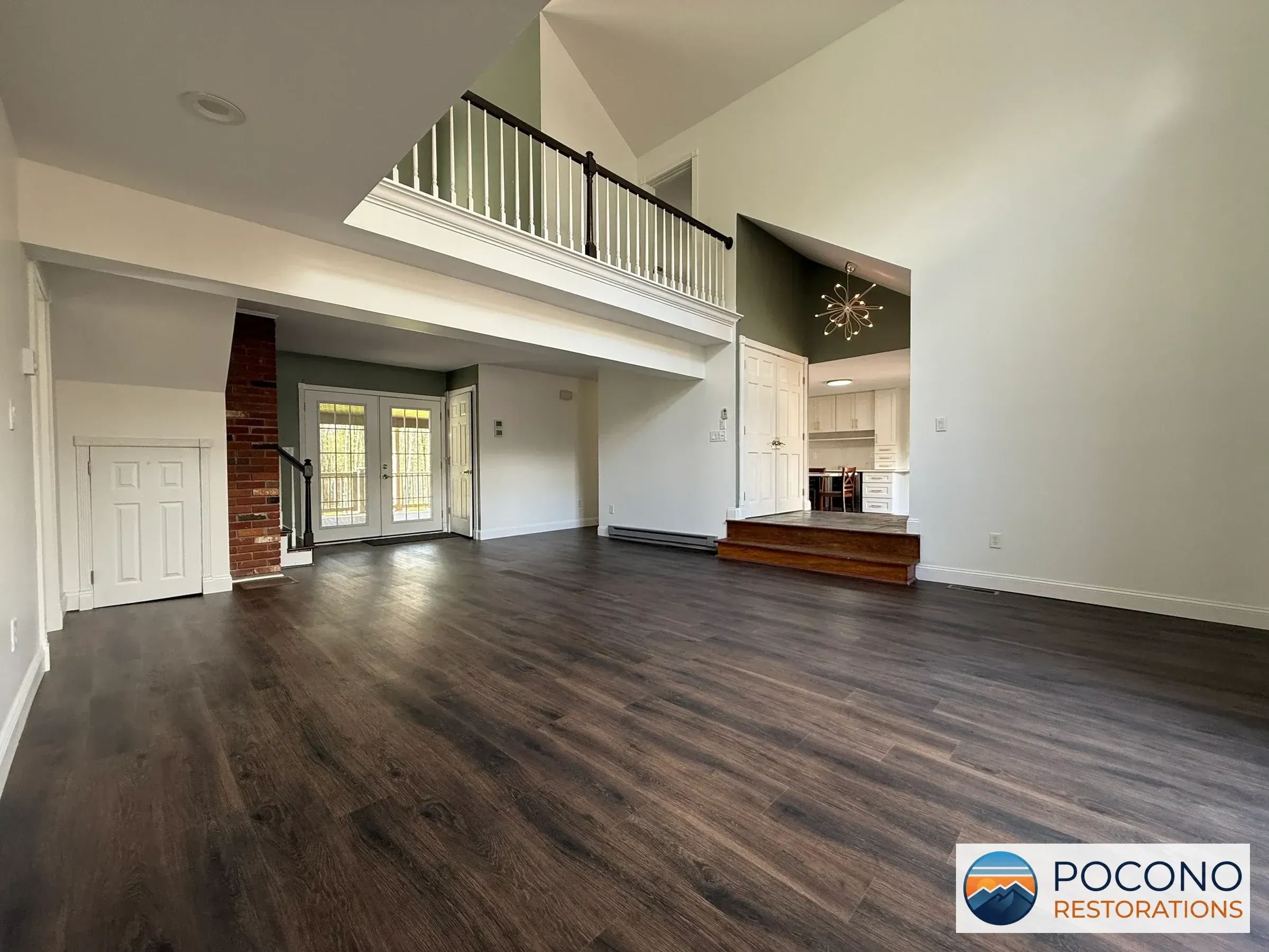 Open living room with dark plank flooring, a vaulted ceiling, a white-railed loft, exposed brick chimney, French doors, and a step up to a white kitchen.
