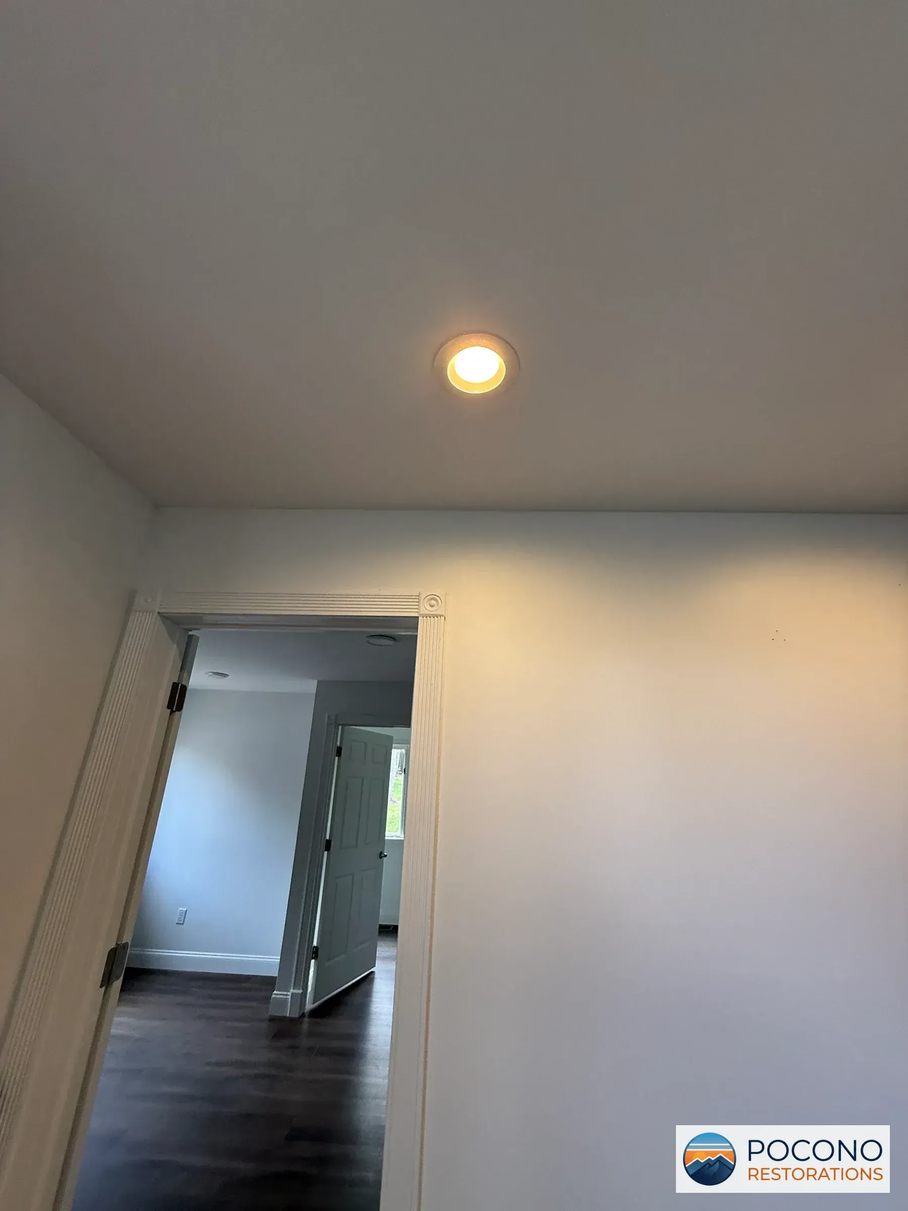 Illuminated recessed ceiling light above a hallway opening into a room with dark wood flooring and an open door.
